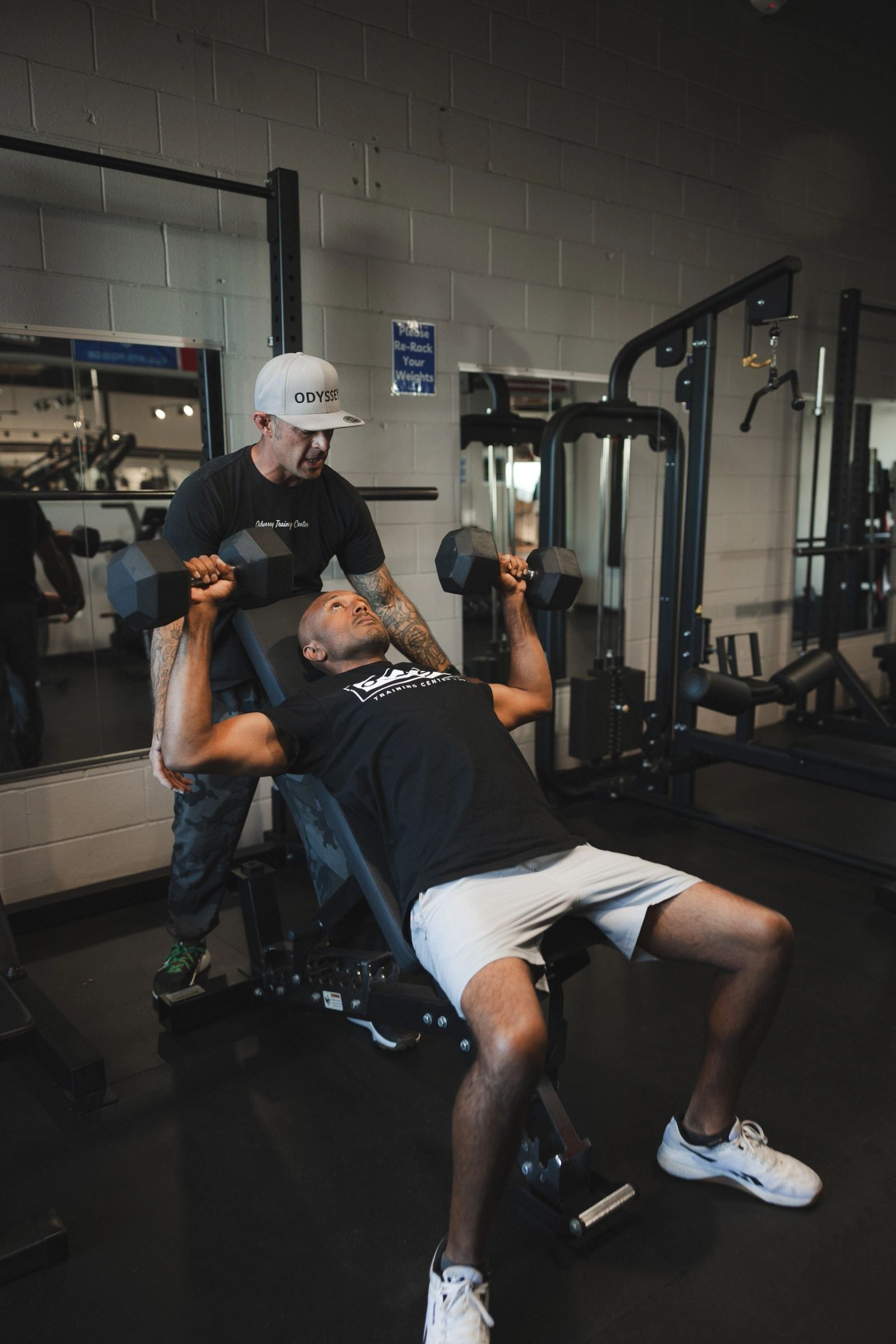 A coach spotting an athlete during a dumbbell press in the Odyssey weight room